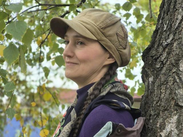 TayaFlavy with a braided ponytail, beige cap, purple shirt, and patterned scarf, smiling near a tree, her ass slightly visible, green leaves in the background.