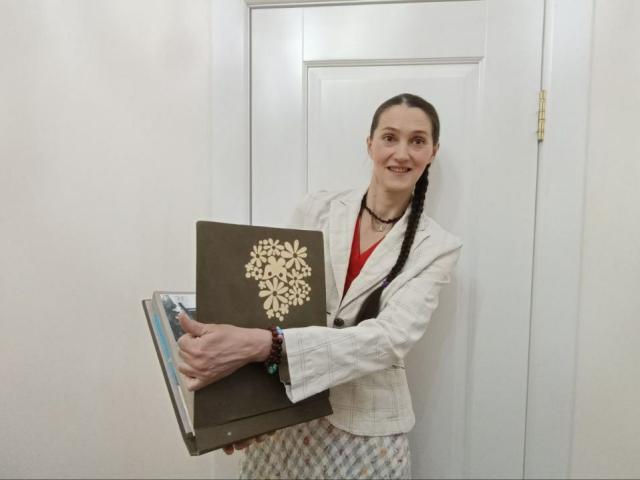 TayaFlavy se tient debout, tenant un livre brun avec une couverture à motifs floraux, portant un blazer blanc, un haut rouge et une jupe à carreaux, avec une tresse et un collier, et elle sourit..