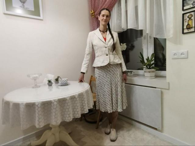 TayaFlavy with small tits, wearing a white blazer and patterned skirt, standing by a lace-covered table, smiling, in a room with a window and potted plant.