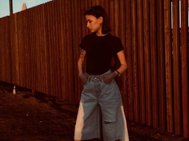 AngelaXSmile stands against a wooden fence, wearing a black t-shirt and high-waisted, loose, light blue jeans with hands in pockets, tattoo on right arm, dark hair.