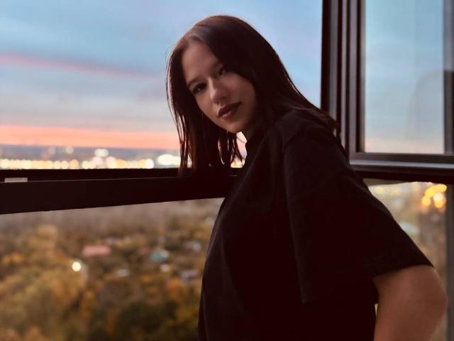 AngelaXSmile with dark red hair, black shirt, standing by a window, looking over shoulder, dusk sky and city lights in background, soft light on her face, slight smile.