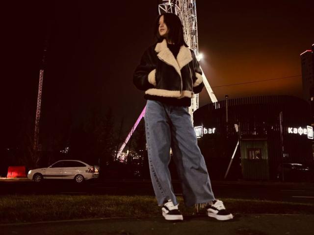 AngelaXSmile in a black puffy jacket with white fur, high-waisted blue jeans, and white Adidas sneakers, standing on a grassy sidewalk at night.