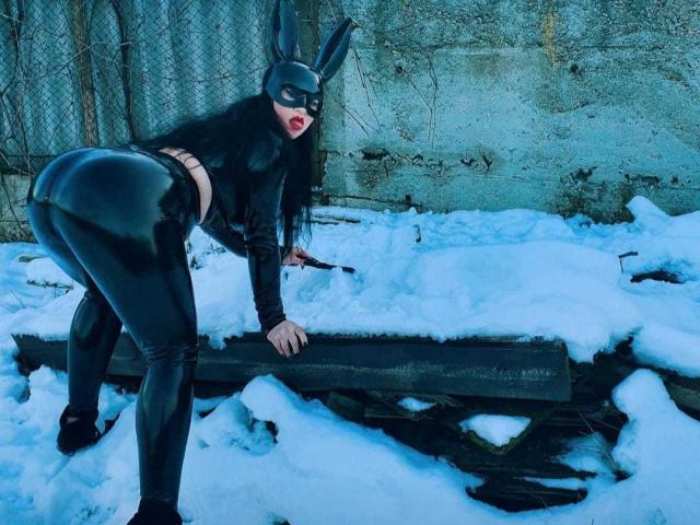 MirayDesire in un outfit di lattice nero, con una maschera a forma di orecchie da coniglio, piegata su una trave di legno in un cortile innevato, che mostra il suo sedere lucido, tenendo una frusta, labbra rosse, capelli scuri..