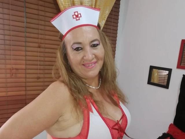 SweetBigCathy in a sexy nurse outfit with red and white lingerie, showing her tits, wearing a nurse cap with a cross, smiling, pearl necklace, and pink lipstick.