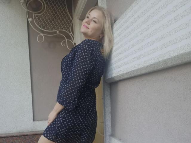 AlliRain with blonde hair, wearing a navy blue dress with white patterns, leans against a wall, smiling, showing her ass slightly, standing near a door with a decorative window.