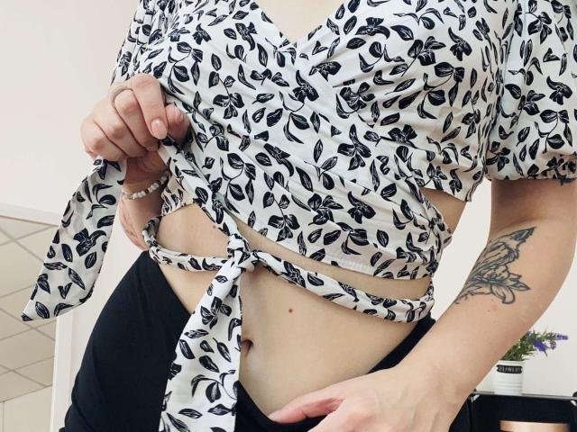 KarKutie lifting her black and white floral top, exposing her small tits and a tattoo on her right arm. She's wearing black pants and a white bracelet.