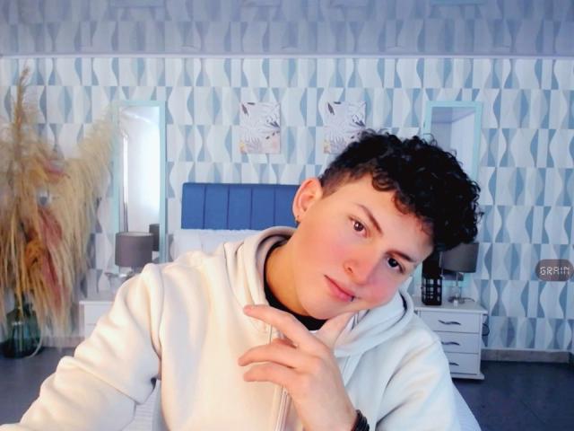 AxelSavage mit lockigem, schwarzem Haar, heller Haut und einem süßen Gesicht, trägt eine weiße Kapuzenjacke, stützt sein Kinn mit dem Finger ab und blickt mit einem leichten Lächeln in die Kamera..