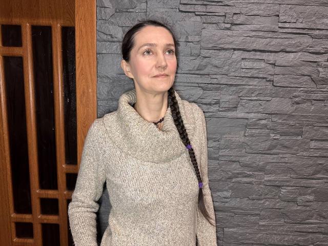 TayaFlavy with a braided ponytail, wearing a grey turtleneck sweater, standing against a stone wall, wooden door with vertical panels on the left. Her tits are small.