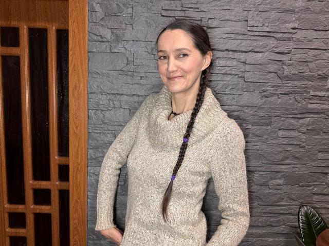 TayaFlavy has a smile, brown braided hair, and is wearing a gray, cabled knit sweater. She's standing against a textured gray wall with a wooden door.