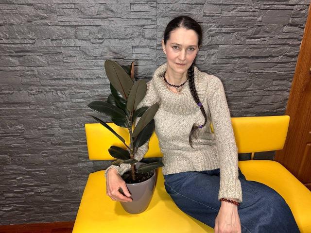 TayaFlavy, with pigtails, sits on a yellow chair, wearing a beige sweater and blue jeans, holding a green plant in a gray pot, against a textured gray wall.
