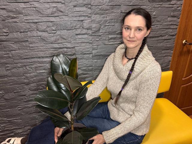 TayaFlavy, with dark braids, sits on a yellow chair, wearing a fluffy gray sweater, holding a green plant, and smiling softly. Her ass is slightly visible in blue jeans.