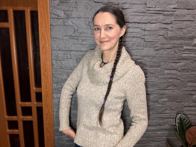 TayaFlavy with long brown braid, smiling, wearing a gray knit cowl-neck sweater, standing against a textured gray wall, hands in pockets, wooden door on the left.