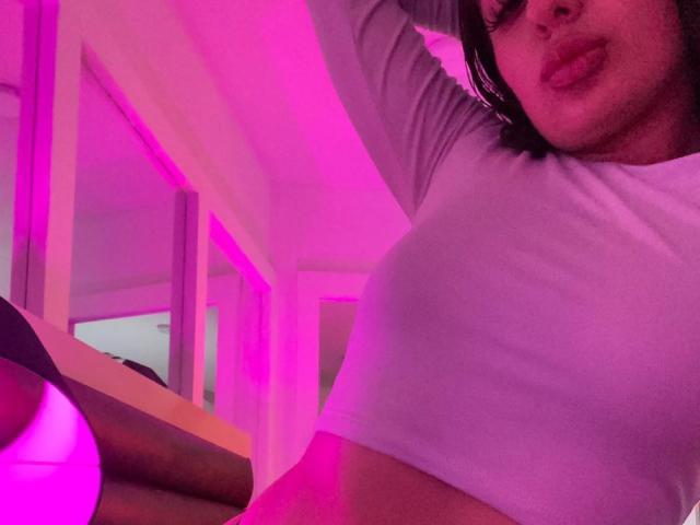 MelissaMora with pouty lips, wearing a white long-sleeve crop top, pink-lit room, raised arm, her tits slightly visible, her face partially in frame, dark hair.