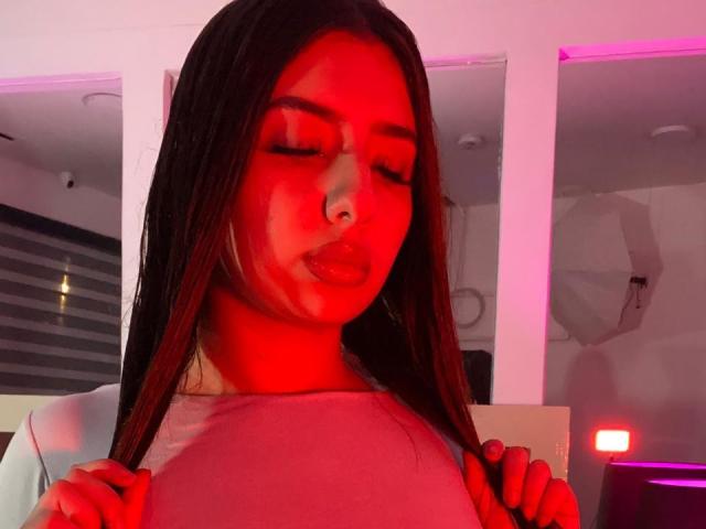 MelissaMora with red light on face, long straight brown hair, closed eyes, pouty lips, holding shirt, nose piercing, pinkish shirt, white background, hands on shirt, subtle smile.