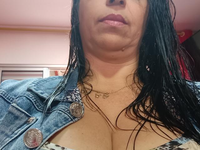 Dede69 con capelli neri e umidi, indossa una giacca di jeans e una maglietta a stampa leopardo, mostrando un seno abbondante e una collana, labbra rosa, una cicatrice leggermente visibile sulla guancia destra..