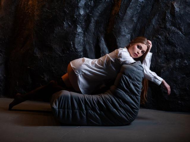 LaraPika with long brown hair, white dress, black boots, and gray quilted chair, lying on floor, arm over chair, looking at camera, dark textured wall in background.