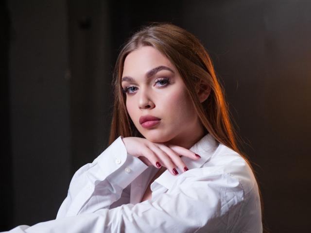 LaraPika with long brown hair, wearing a white shirt, has her chin on her hand, looking serious with red lipstick and manicured nails, in a dark, moody setting.