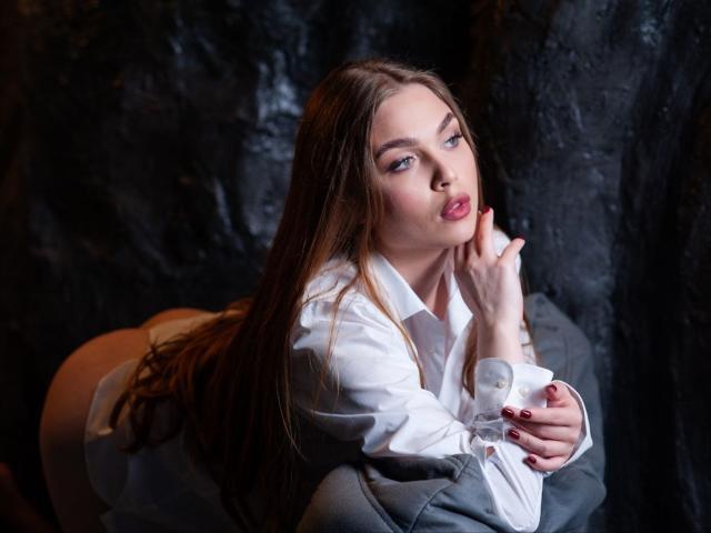 LaraPika with long brown hair, white shirt, and red nails, sucking her finger, looking at the camera, kneeling with her ass up, on a dark textured surface.