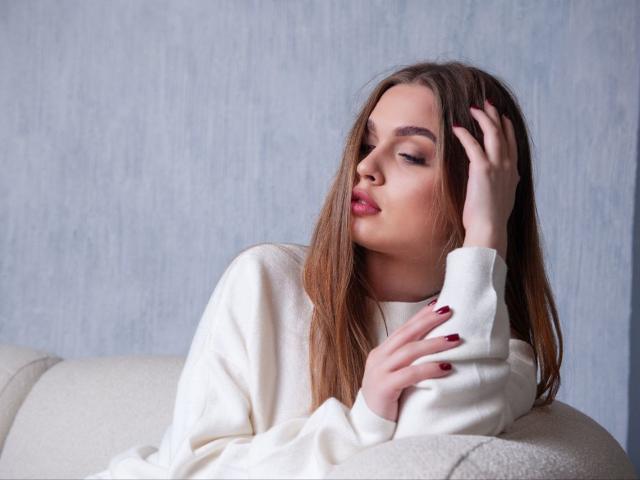 LaraPika with long brown hair, eyes closed, wearing a white sweater, red nail polish, hand in hair, sitting on a white couch, looking slightly bored or pensive.