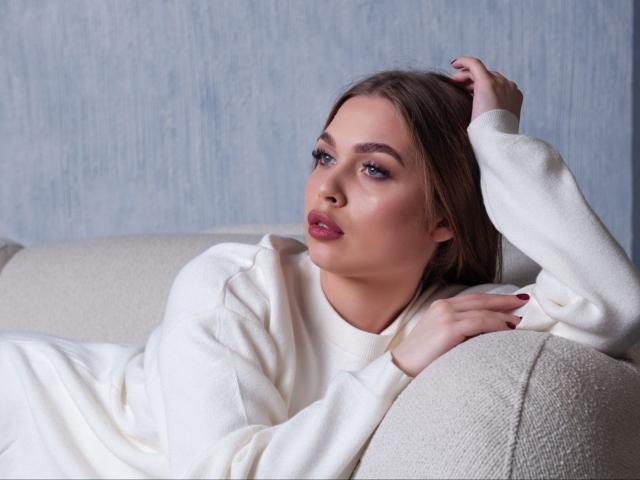 LaraPika with light skin, brown hair, and red lipstick, wearing a white sweater, lying on a beige couch, hand in hair, looking slightly to the side.