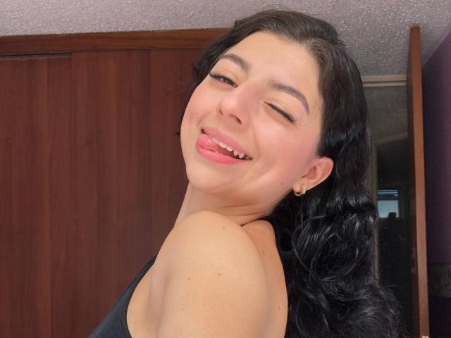 AnntonellaWatson with black wavy hair, sticking out her pink tongue, winking, wearing a black tank top, showing her shoulder and small earrings, smiling.