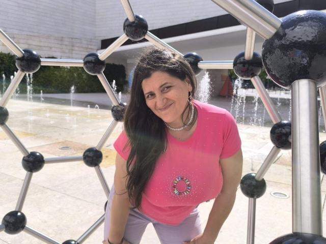 BlissSophia, avec ses longs cheveux noirs, sa chemise rose, son collier de perles et son pantalon violet, est appuyée contre une structure métallique, souriante, avec le soleil sur son visage, et des fontaines en arrière-plan..