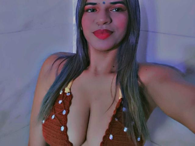 KairaDayamai with long grayish-brown hair, brown eyes, and red lipstick, wearing a brown, deep cleavage, crochet top, showing her big, rounded boobs and a nose ring.