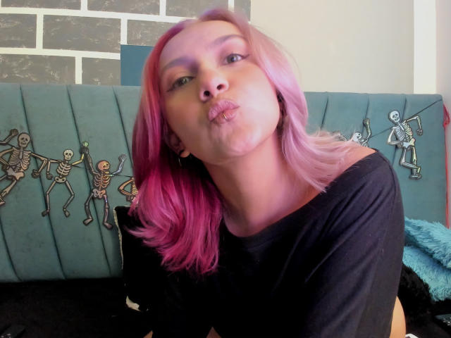 AlterEgoSecond with pink hair, black shirt, pouty lips, and a cute face, sitting on a blue couch with skeleton decorations. She's making a kissing face.