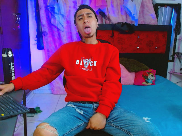 Abnedarm is a dude with brown skin and a beanie, tongue out, wearing a red sweatshirt and ripped jeans, sitting on a bed with a blue blanket.