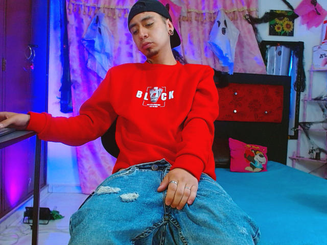 Abnedarm wearing a black backward cap, red sweatshirt, and blue jeans with a rip, sitting on a bed, looking down with a slight pout, hands resting on knees.