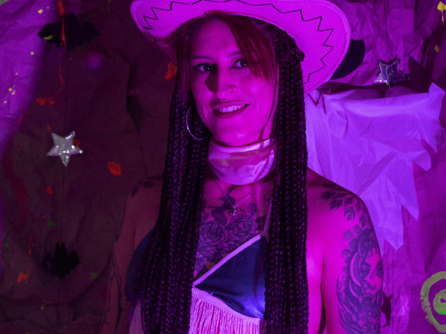 SofiFw with long braids, white cowboy hat, pink choker, tattooed chest and arm, smiling, wearing a blue and white top, purple lighting, visible nose ring, and hoop earrings.