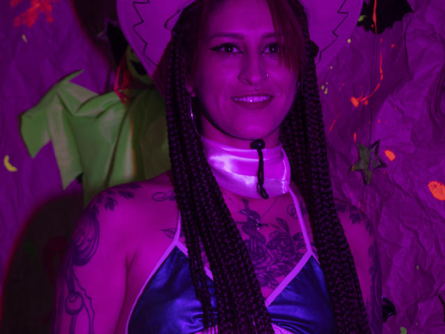 SofiFw with purple-tinted skin, wearing a shiny blue top, white hat, braided hair, tattoos, nose ring, smiling, and a neon pink choker. Boobs, pussy, and ass not visible.