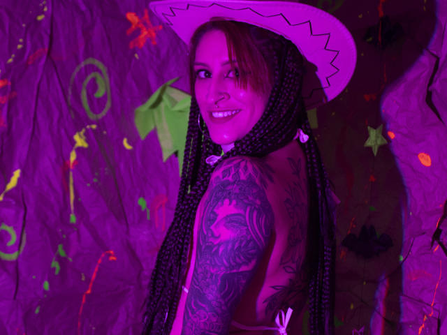 SofiFw with purple-lit face, long braids, white cowboy hat, tattooed arm, and smiling, showing her tits, in a colorful, neon background. She's looking over her shoulder.