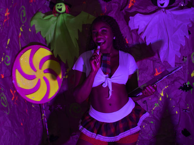 LeleFw with dark skin, curly hair, and small tits, wearing a white crop top, plaid skirt, and red stockings, holds a lollipop, standing in front of Halloween decorations.