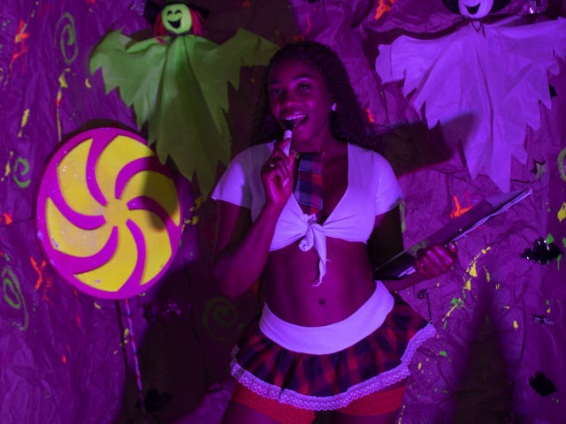 LeleFw, dark-skinned woman with curly hair, in a white tied-up top, red plaid skirt, and orange stockings, holding a microphone, surrounded by Halloween decorations, including a swirl candy.