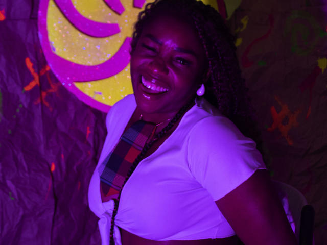 LeleFw, smiling with one eye closed, has dark skin, curly hair, white top, plaid shirt, and white earrings, showing her boobs, under purple and yellow graffiti.
