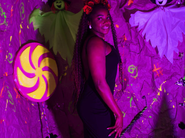 LeleFw in a black dress, smiling, standing with curly hair, purple lighting, Halloween decorations, and a giant yellow swirl candy on the left. She's holding her ass with one hand.