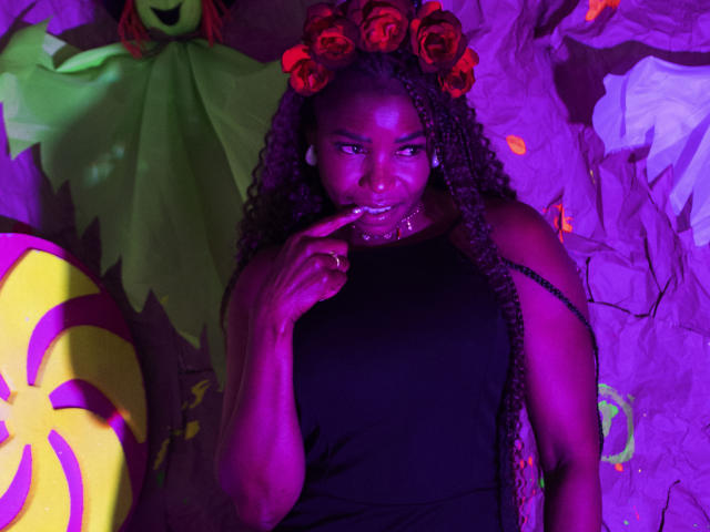LeleFw with dark skin, curly braids, wearing a black sleeveless top, red rose headpiece, and purple lighting, has a finger to their lips, looking seductive.