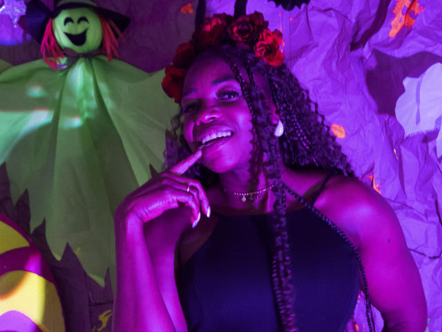 LeleFw, dark-skinned with long braids, wearing a red flower crown, black dress, and necklace, smiling, with finger to mouth, purple lighting, Halloween decoration in background.