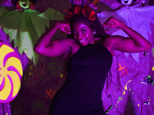 LeleFw, dark-skinned, muscular, smiling, wearing black dress, arms flexed, curly hair with red roses, purple and pink lighting, standing in front of Halloween decorations, showing strong arms.