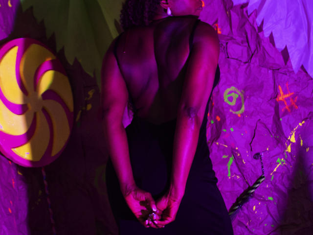 LeleFw, back to camera, topless, black pants, muscular, dark skin, tattoo on right arm, hands clasped, purple and green light, large yellow and pink swirl on left.
