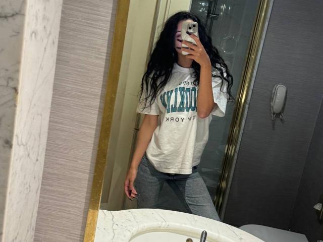 Mirror selfie of ShadowDance with long black hair, white t-shirt, gray pants, holding phone, covering face, standing in bathroom, showing ass, wearing loose clothes, looking at phone.