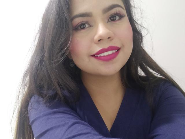 LilyRousee with long dark hair, pink lipstick, and purple eyeshadow, wearing a dark blue top, smiling at the camera. She has pink cheeks and small earrings.