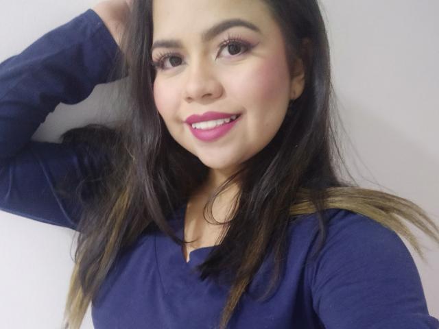 LilyRousee with long brown hair, blue shirt, pink lipstick, smiling, hand in hair, dark eyebrows, long eyelashes, light skin, subtle blush, looking at camera, cute, sexy face.
