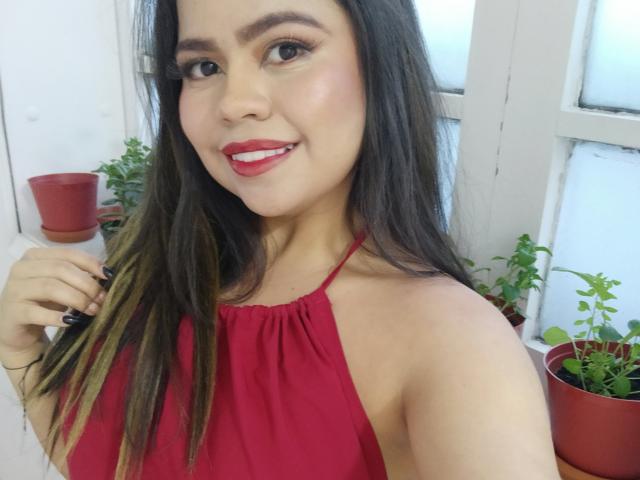 LilyRousee has long, straight brown hair with subtle blonde highlights. She wears a red halter top, revealing her shoulders. Her red lipstick matches her top, and she has a slight smile. She has light skin and dark eyebrows. Her right hand, with black nail polish, touches her hair. The image has a casual, selfie-style composition. Two potted plants are in the background, one red and one green. The window behind her is slightly blurred. The overall style is bright and candid.