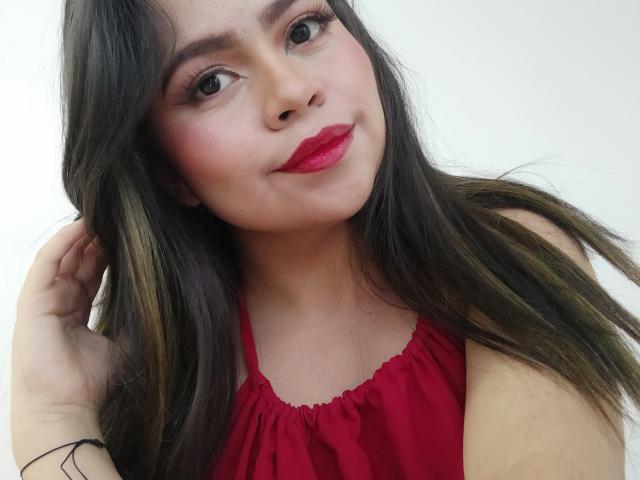 LilyRousee has long brown hair, red lipstick, and a red dress. She's smiling with a hand in her hair, showing off her tits slightly. She's looking cute.