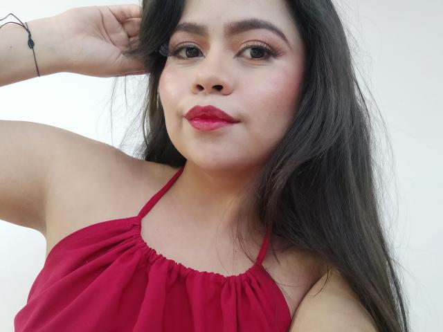 LilyRousee has long black hair, red lipstick, and is wearing a red halter top, with a small black bracelet on her right wrist, looking at the camera.