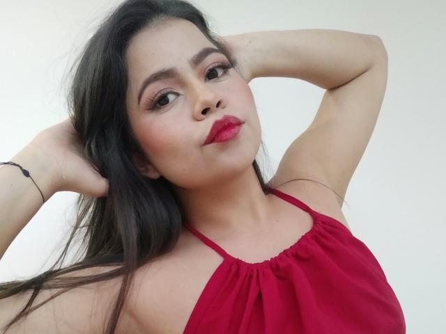 LilyRousee with long black hair, red lipstick, and red halter top, arms raised, looking at camera, soft smile, light skin, subtle makeup, bracelet on right wrist.