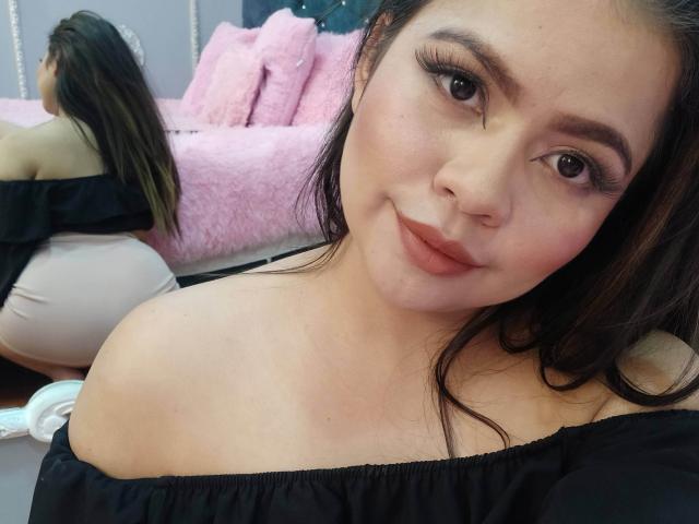 LilyRousee with long brown hair, black off-shoulder top, and pink lips, sitting on floor, showing her ass in mirror reflection, wearing white pants, in bedroom.