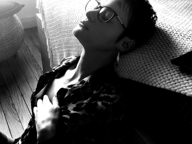 LaPhoenixElo with short dark hair, glasses, and a patterned shirt, lying on a bed, eyes closed, hand on chest, in black-and-white, soft light, looking relaxed.